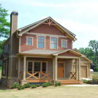 Brown, wooden house with a chimney on the left side. It has a large front porch and is surrounded by many green trees.