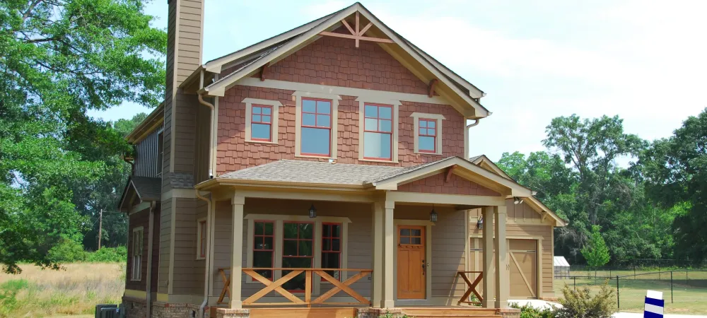 house-with-chimeny Brown, wooden house with a chimney on the left side. It has a large front porch and is surrounded by many green trees.